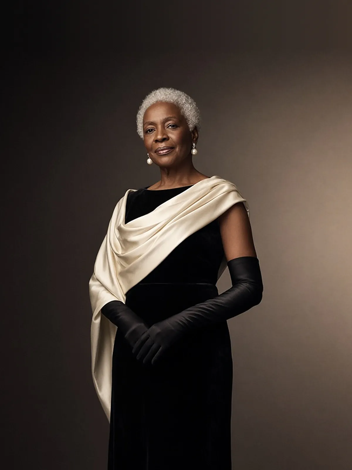 AI campaign art: A silver-haired older Black woman with a warm, knowing smile poses against a dark umber backdrop in a black velvet gown, an ivory satin stole draped over one shoulder, long black opera gloves and pearl drop earrings.